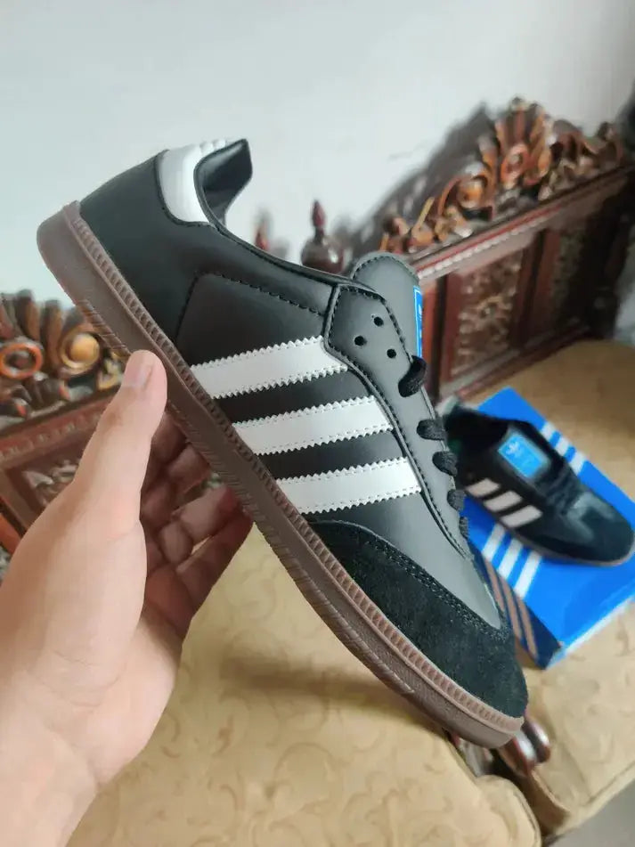 Adidas Samba OG Black sneaker held in hand with original box in background on decorative chair.