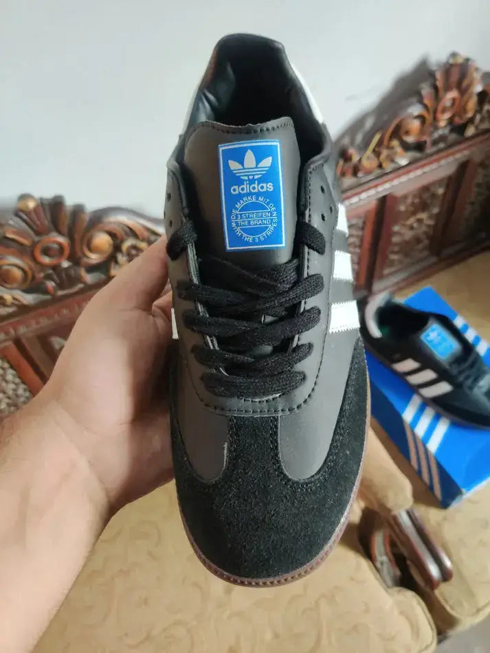 Adidas Samba OG Black sneaker held in hand, showcasing iconic design with blue and white branding.