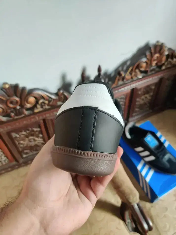 Close-up of Adidas Samba OG Black sneaker with classic design, showcasing iconic style against a decorative backdrop and shoe box.